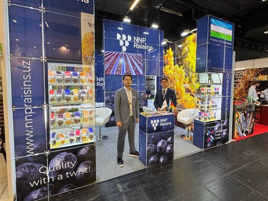 The NNP Raisins stand at Anuga 2025 seen from the aisle, two of the team in front of it, a tall lit cabinet of packed raisins on the left and a Product of Uzbekistan panel on the right.