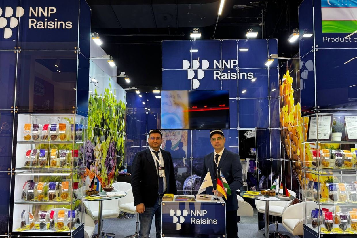 Two members of the NNP Raisins team inside the company's stand at Anuga 2025, the NNP Raisins mark on the back wall and the counter, lit glass cabinets of packed raisins on both sides and the Uzbek flag panel above.