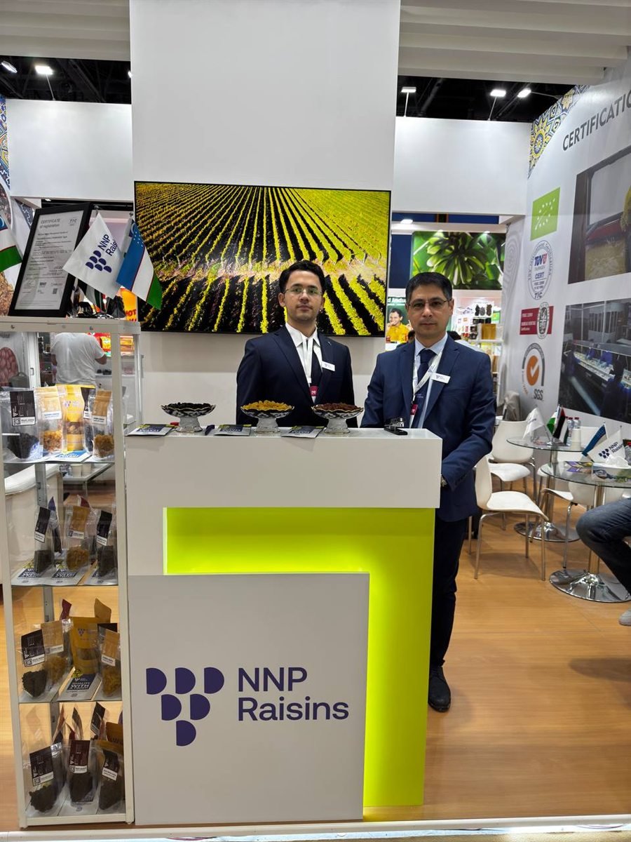 Two of the NNP Raisins team behind the company's counter at Gulfood 2025, dishes of golden and dark raisins on the desk, packed samples on the shelves beside them, a framed certificate standing next to the NNP and Uzbek flags, and aerial footage of the vineyards on the screen above.