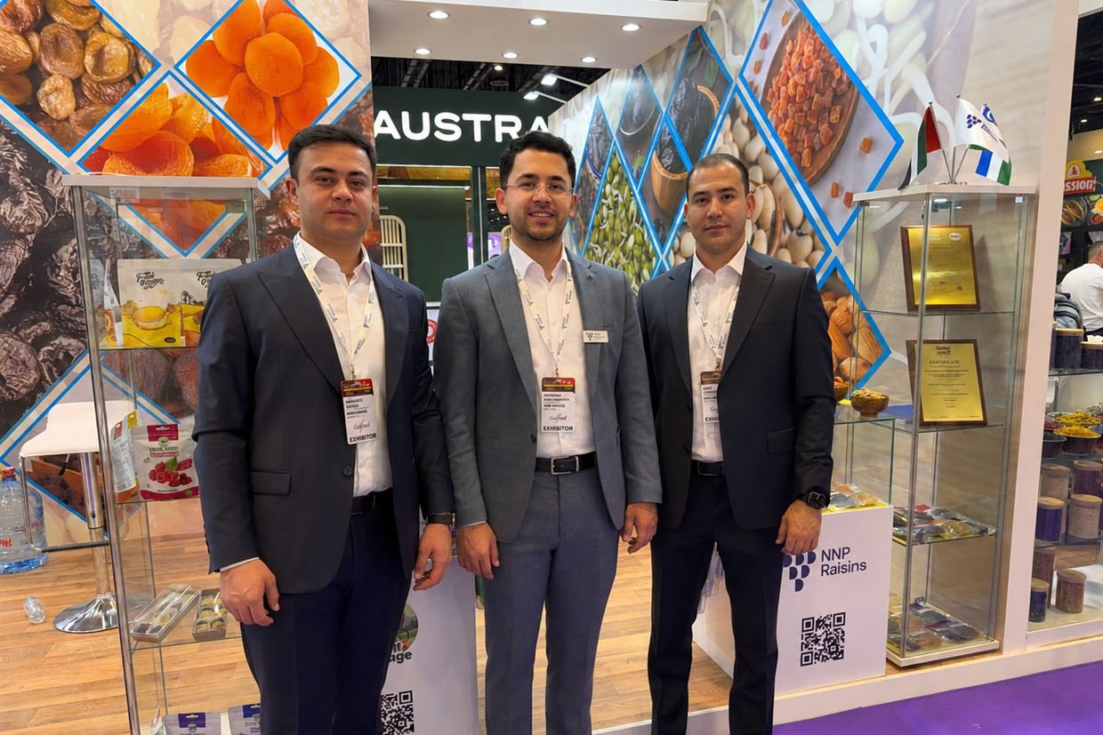Three members of the NNP Raisins team at the company's stand at Gulfood 2026, the NNP Raisins mark and a QR code on the counter behind them, product shelves and a glass display case either side.