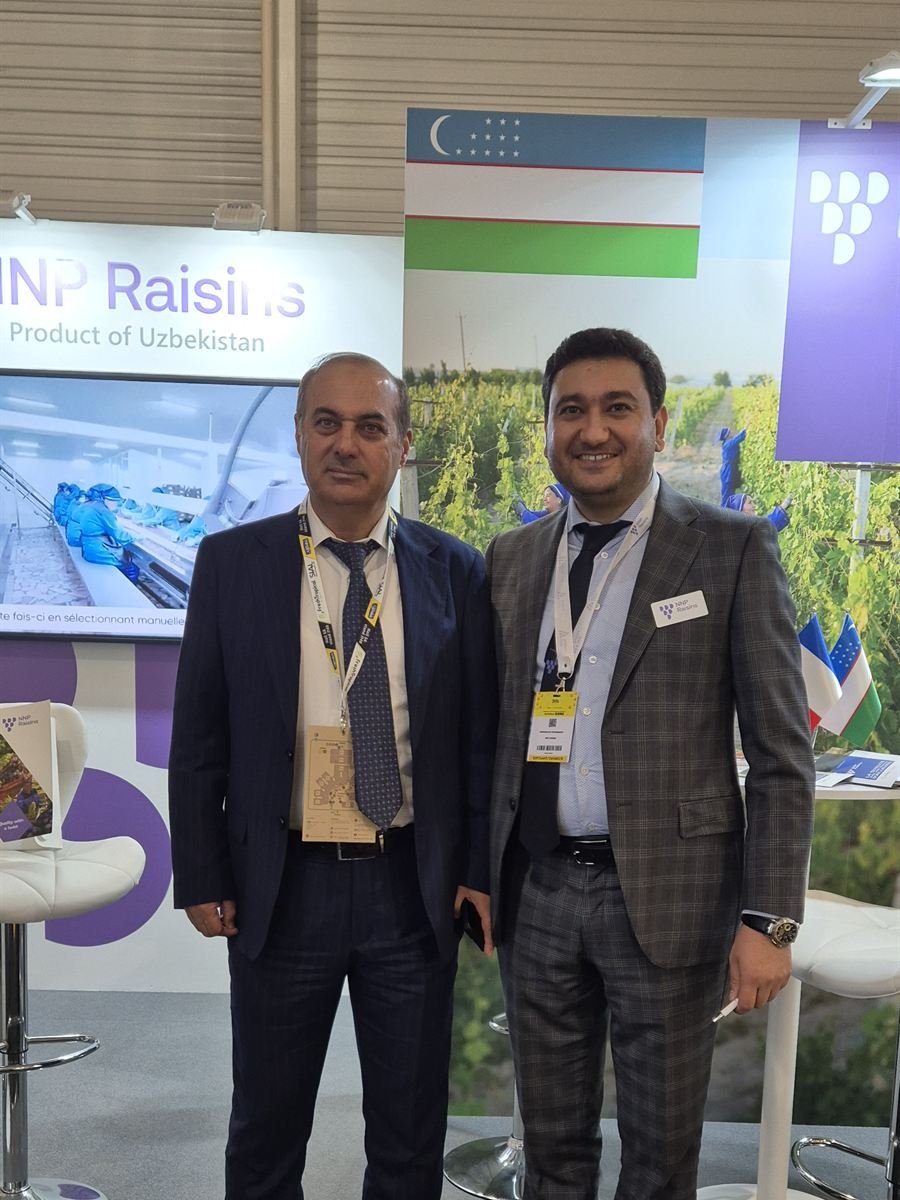 Two of the NNP Raisins team at the stand at SIAL Paris 2024, in front of a Product of Uzbekistan panel and the Uzbek flag, with the sorting-line footage on the screen beside them.