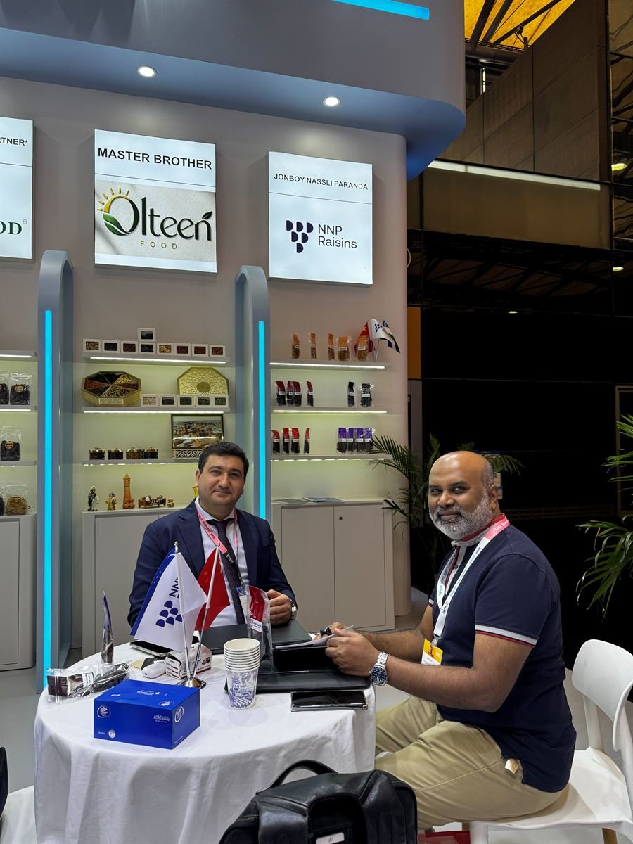 A meeting at the NNP Raisins stand at SIAL Shanghai 2026: two people seated at a round table with samples, a notebook and an NNP Raisins table flag, the product shelves behind them.