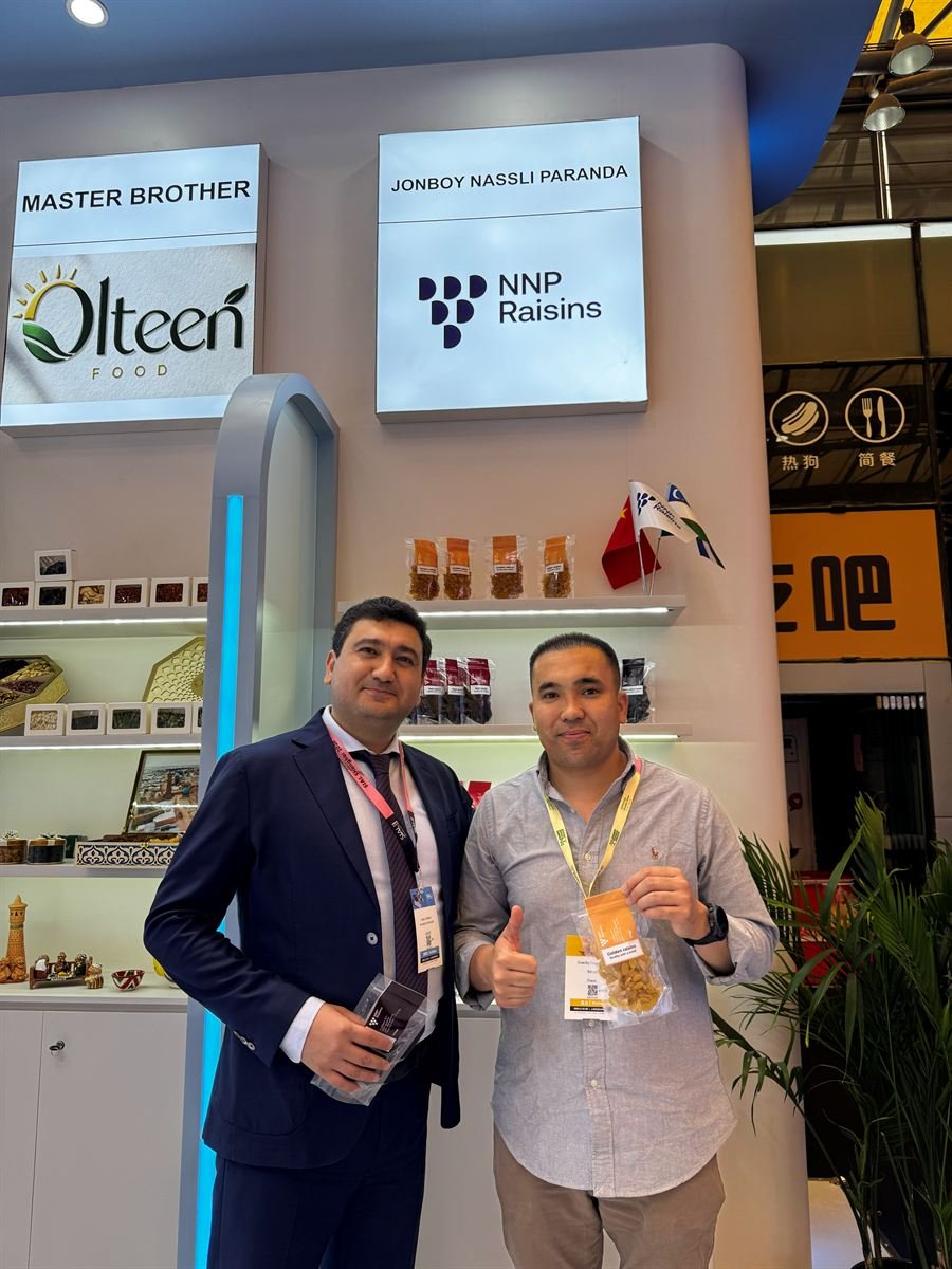 Two members of the NNP Raisins team at the stand at SIAL Shanghai 2026, the illuminated NNP Raisins sign above them, packs of raisins on the shelves behind and the Uzbek and Chinese flags on the counter.